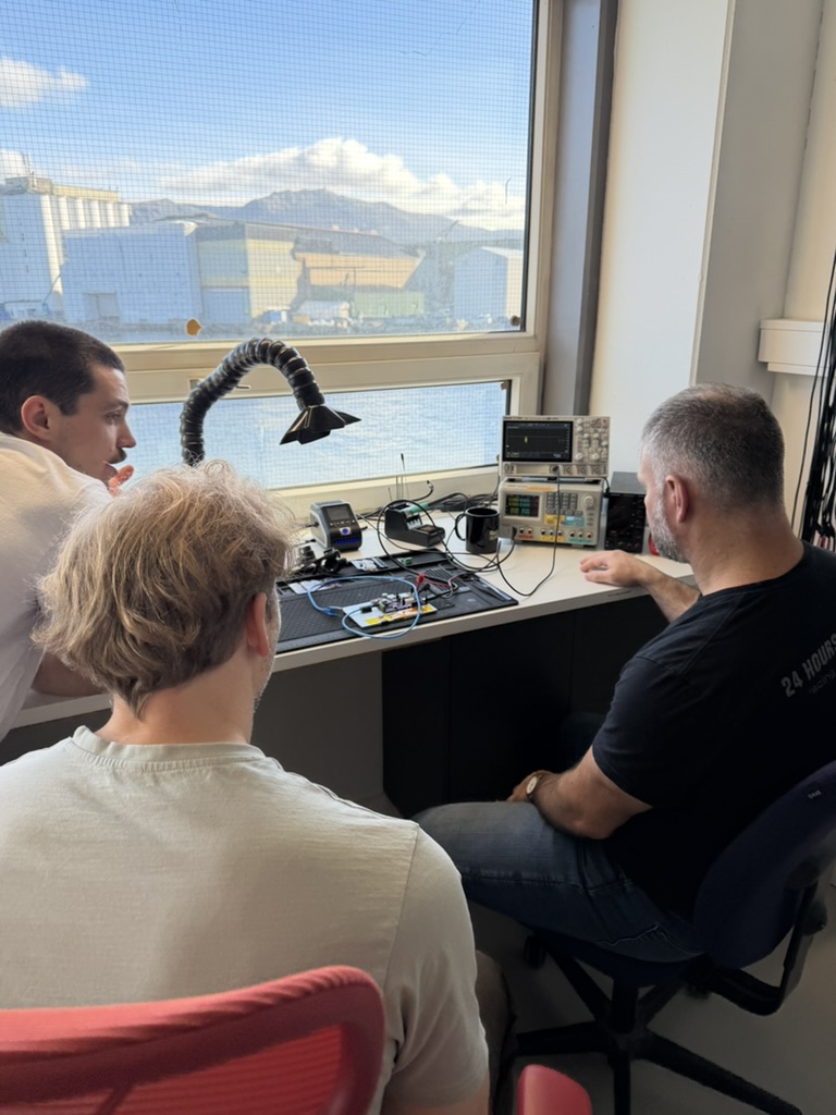 Three team members around the electronics bench by a window over the harbour