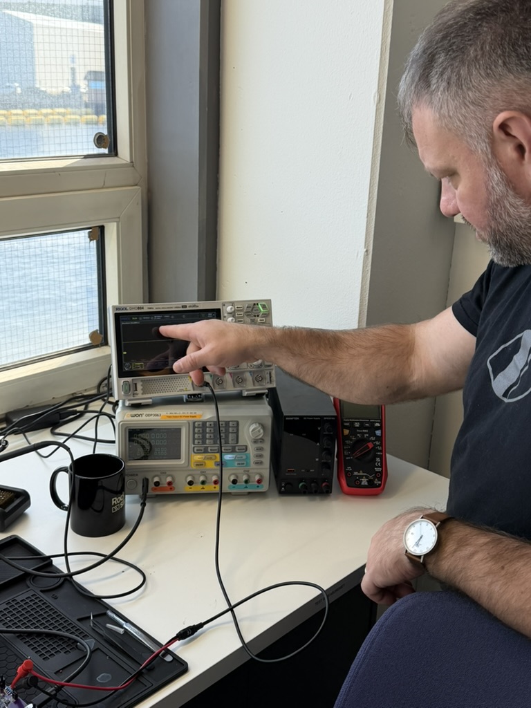 Daniel pointing at an oscilloscope on the electronics bench